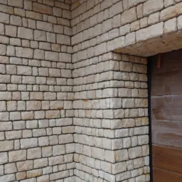 Fragment elewacji budynku wykończony jasnym kamieniem naturalnym o nieregularnym kształcie, widoczny narożnik budynku i fragment drewnianych drzwi.