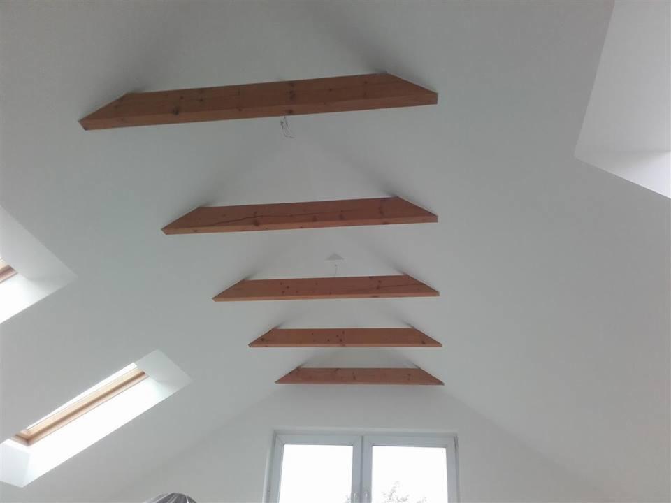 Białe poddasze z ukośnym sufitem i szeregiem drewnianych belek stropowych ułożonych równolegle, okna dachowe i okno pionowe w dolnej części kadru.