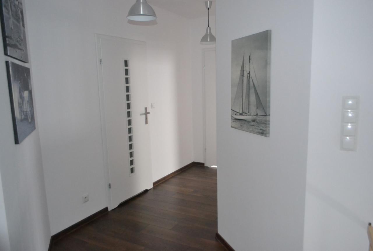 Wnętrze przedpokoju po remoncie: białe ściany, ciemna drewniana podłoga, nowoczesne oświetlenie, drzwi z dekoracyjnymi wgłębieniami i obraz z motywem żaglówki na ścianie.