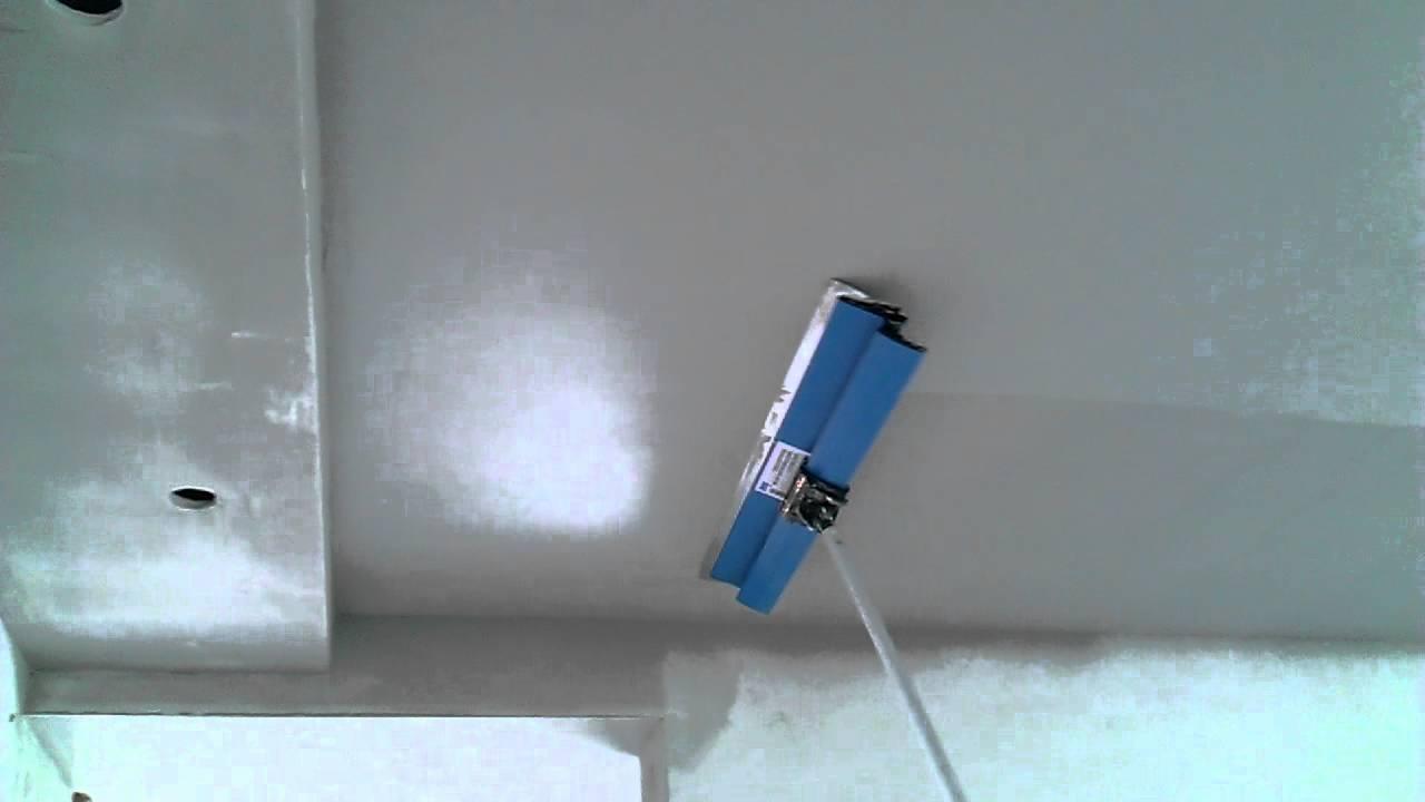 Aplikacja gładzi na suficie za pomocą szpachli z długim uchwytem, widoczne nierówności na powierzchni przed ostatecznym wygładzeniem.