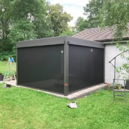 Nowoczesna, szara pergola tarasowa z czarnymi roletami zewnętrznymi, widok z zewnątrz na trawnik i dom, drabina i narzędzia obok sugerują montaż w toku.