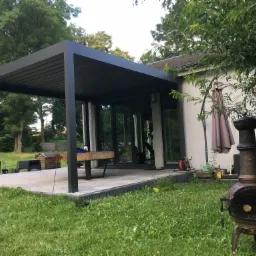 Nowoczesna, czarna pergola z żaluzjowym dachem przylegająca do domu z dużymi, przesuwnymi oknami, otoczona zielonym trawnikiem i drzewami.