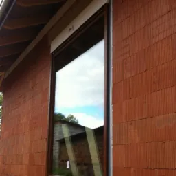 Brązowe okno z roletą zewnętrzną zamontowane w ścianie z czerwonej cegły kratówki, widoczne odbicie budynku w szybie.