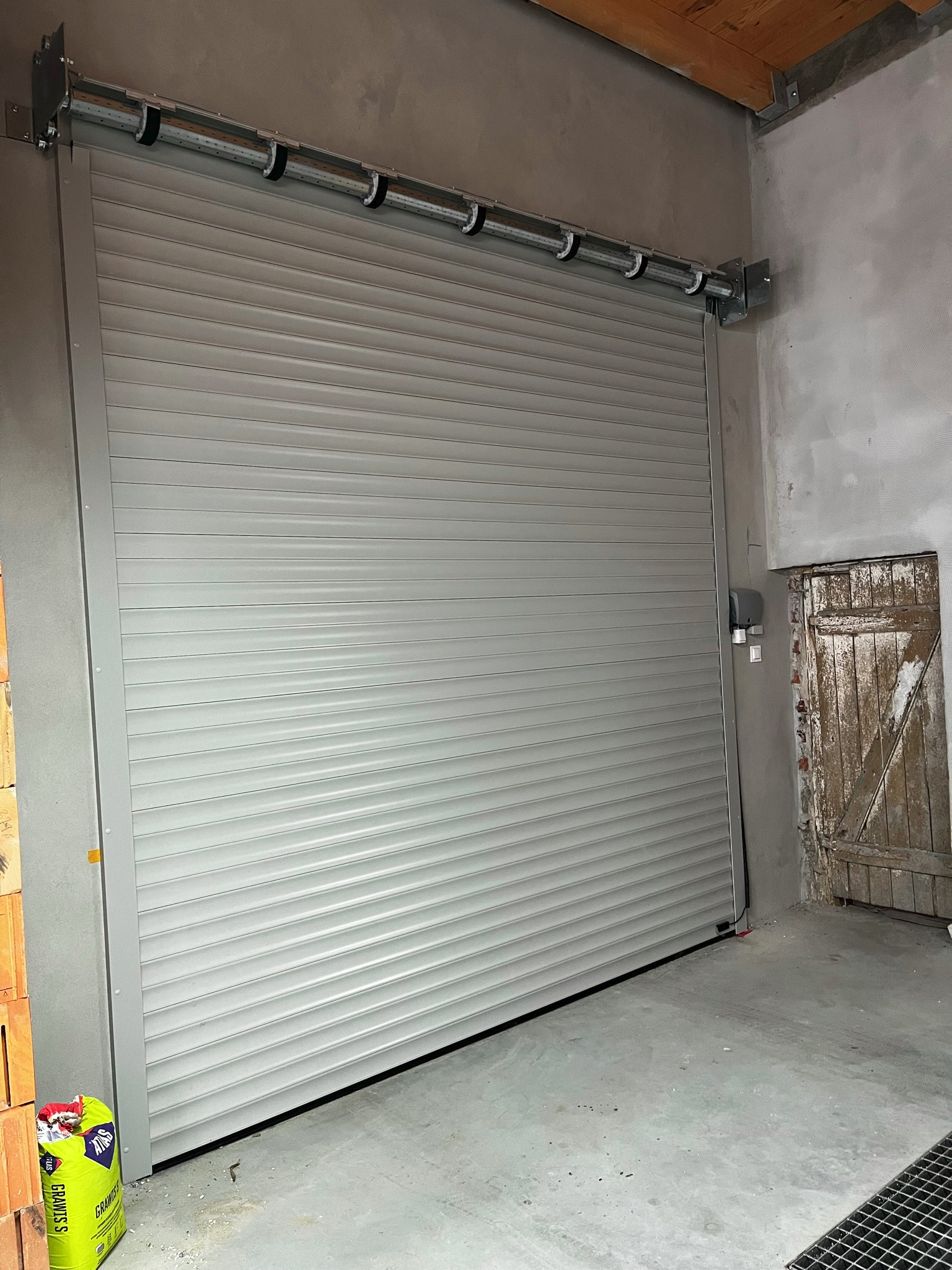 Szara brama rolowana garażowa z napędem elektrycznym, widoczne prowadnice i mechanizm zwijania, obok stara drewniana futryna drzwi i worek z zaprawą murarską.