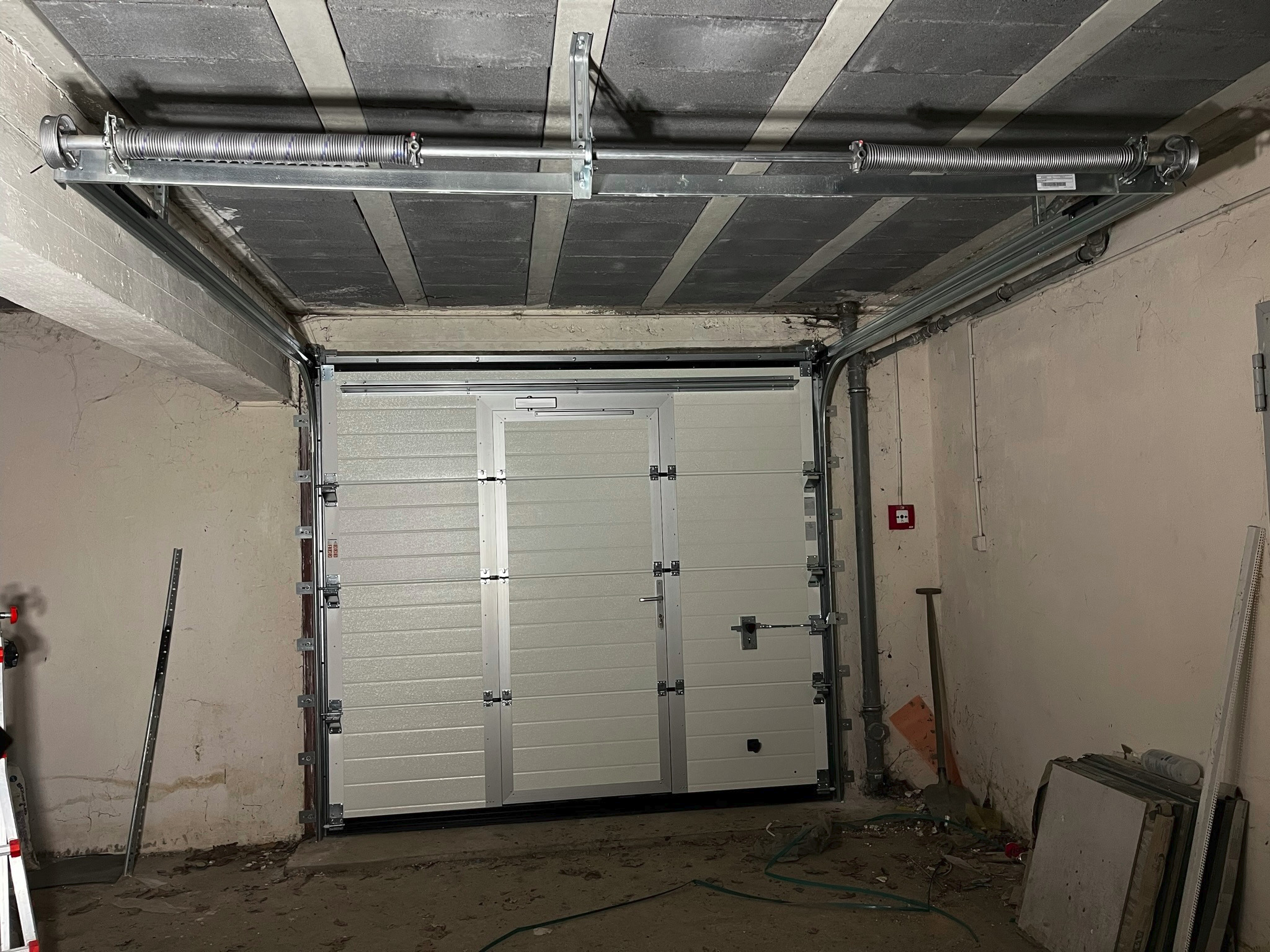 Zamknięta segmentowa brama garażowa w surowym garażu z widocznym mechanizmem sprężynowym pod betonowym sufitem.