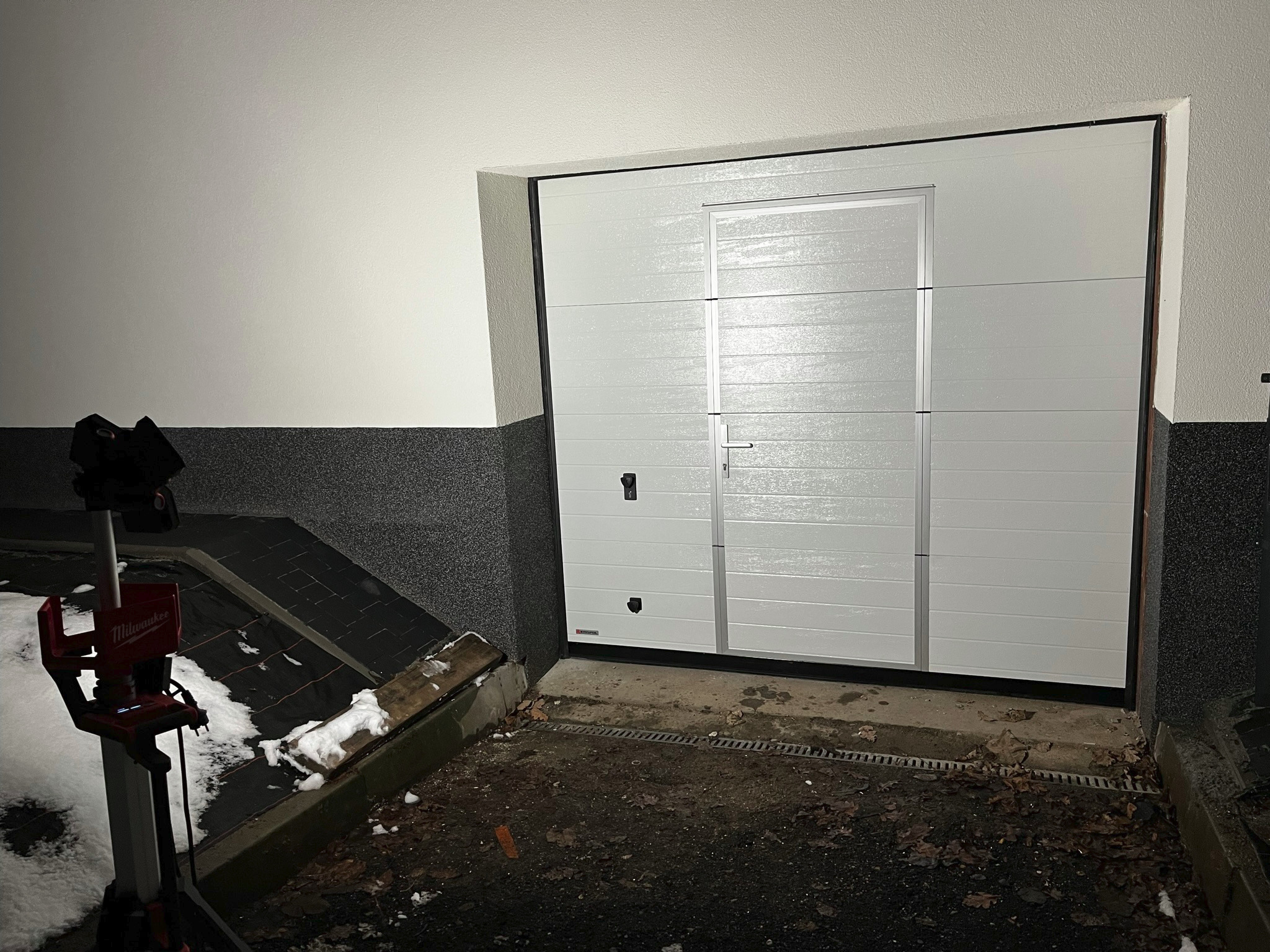 Zamknięta, segmentowa brama garażowa w kolorze białym, z drzwiami serwisowymi po prawej stronie, oświetlona lampą roboczą Milwaukee, na tle ściany z szarą, ziarnistą fakturą.
