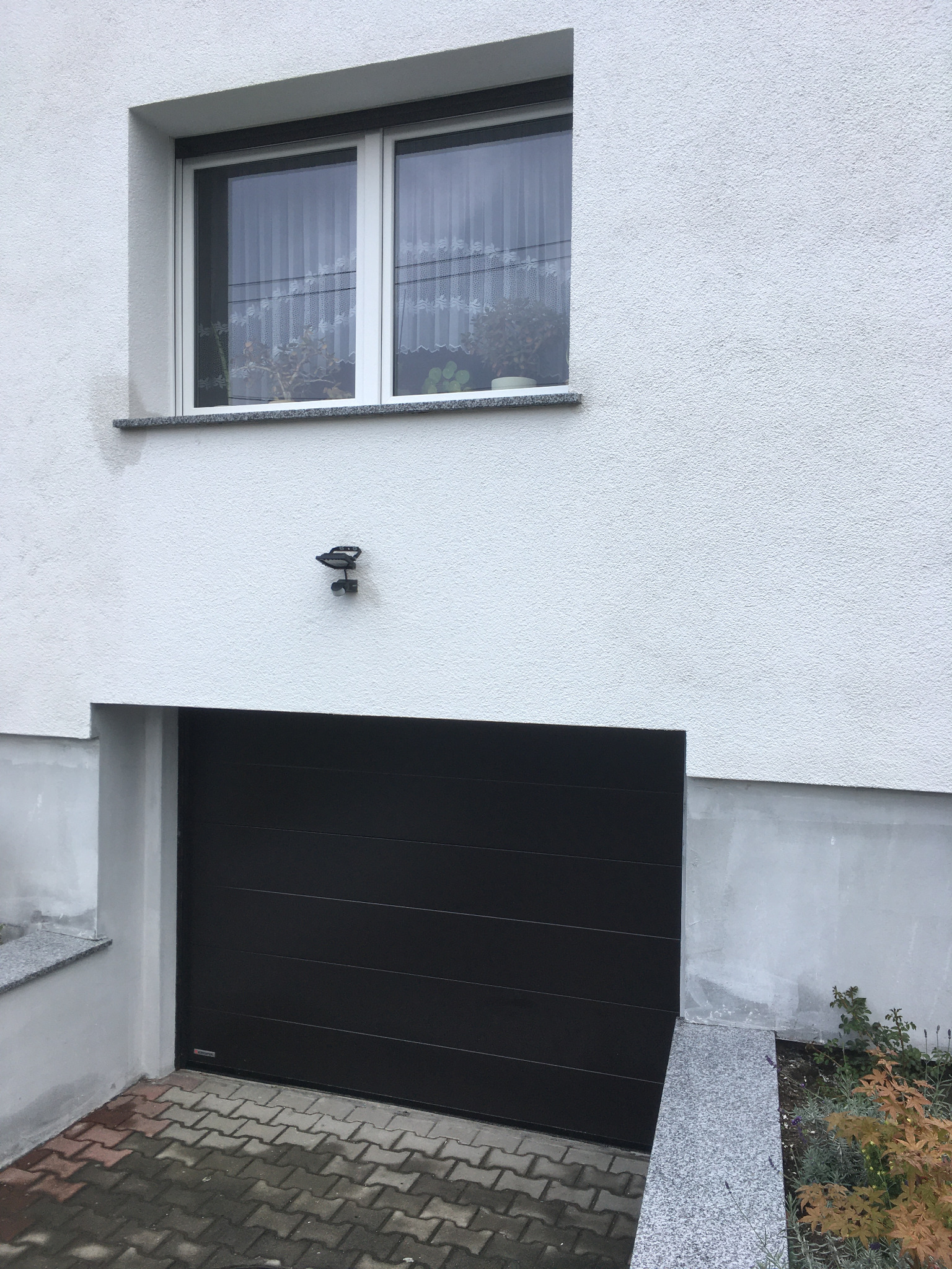 Ciemna brama garażowa segmentowa, widok z zewnątrz, z oknem PCV powyżej i lampą zewnętrzną na elewacji z tynkiem strukturalnym.