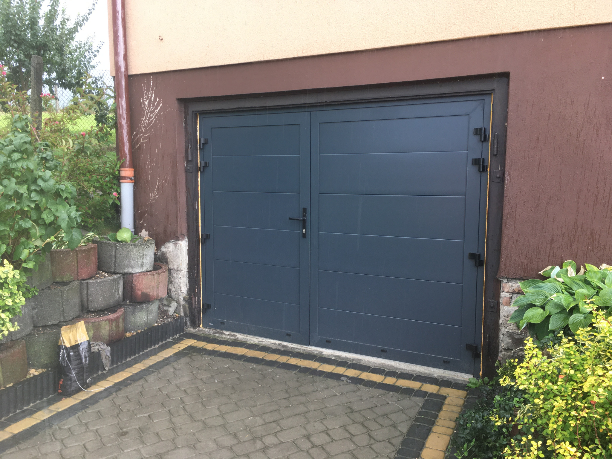 Szara, dwuskrzydłowa brama garażowa z poziomymi panelami, zamontowana w brązowym budynku, z widocznym fragmentem kostki brukowej i zieleni po bokach.