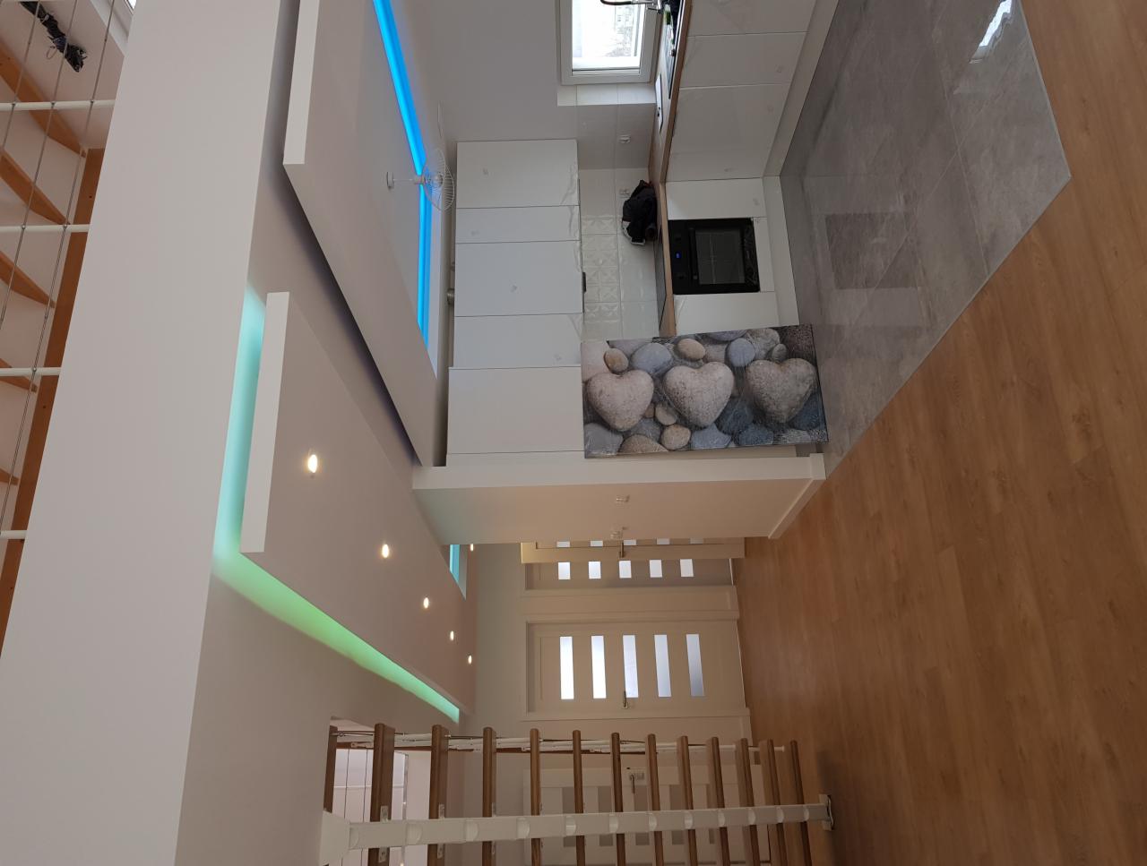 Nowoczesna kuchnia z białymi meblami, szarą podłogą i dekoracyjnym panelem z motywem kamieni w kształcie serc, oświetlenie LED w suficie, widok z góry na otwarte drzwi.