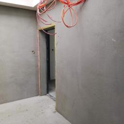 Ravbud - Surowe, szare ściany po nałożeniu tynku maszynowego, widoczne wystające kable elektryczne i ościeżnica drzwiowa zabezpieczona folią, w tle sufit z oknem dachowym.