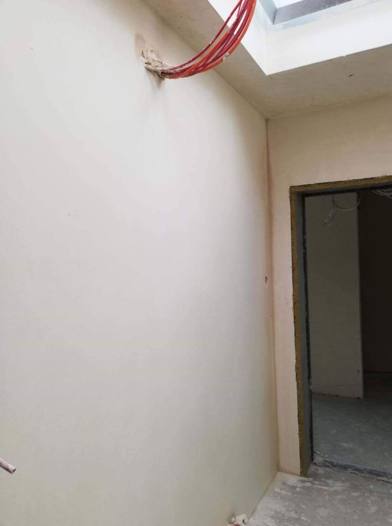 Świeżo otynkowana ściana z wystającymi przewodami elektrycznymi w rogu, przygotowana do dalszych prac wykończeniowych, widoczny fragment sufitu z oknem dachowym i surowy otwór drzwiowy.