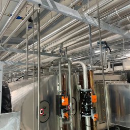 Grand - Kompleksowa instalacja rur wentylacyjnych i hydraulicznych w budynku przemysłowym, widoczne liczne rury ze stali nierdzewnej, zawory, czujniki oraz elementy mocujące.