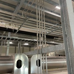 Grand - Metalowe rury wentylacyjne i hydrauliczne w hali przemysłowej, widoczne elementy mocujące i podwieszana konstrukcja sufitowa.