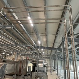 Grand - Widok na system rur i urządzeń wentylacyjnych w hali przemysłowej, z widocznymi elementami mocującymi i niewielką postacią ludzką w tle.