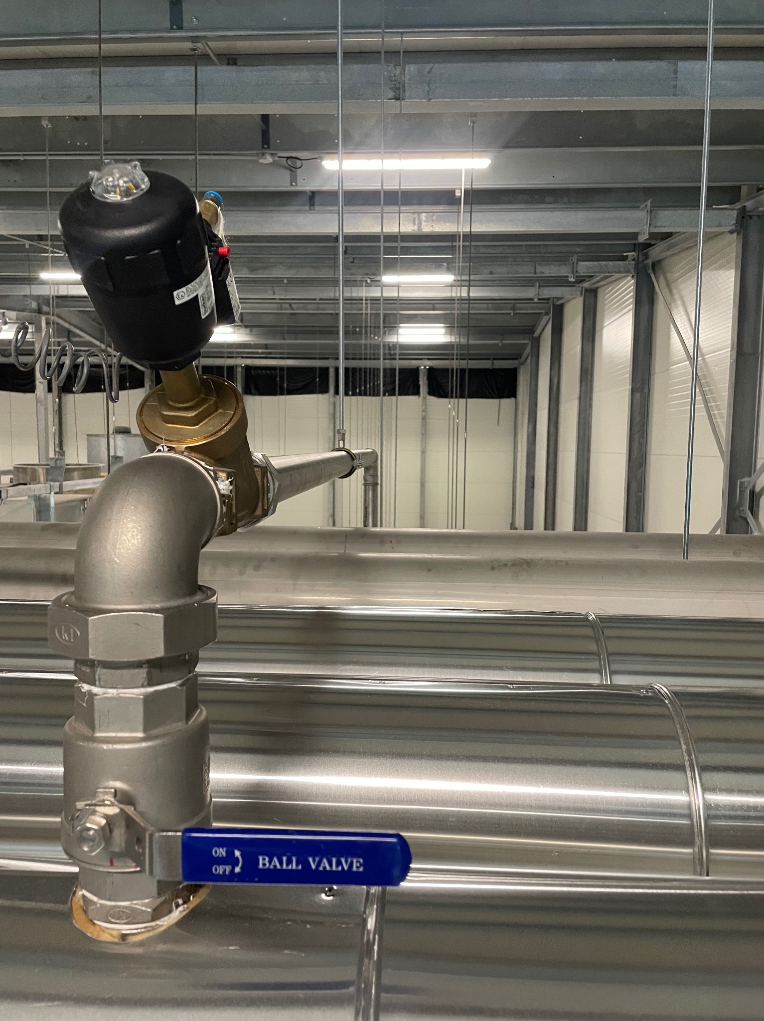 Zamknięty zawór kulowy z niebieską dźwignią na instalacji rurociągowej w hali przemysłowej, widoczny filtr na rurze pionowej i metalowa konstrukcja pod sufitem.