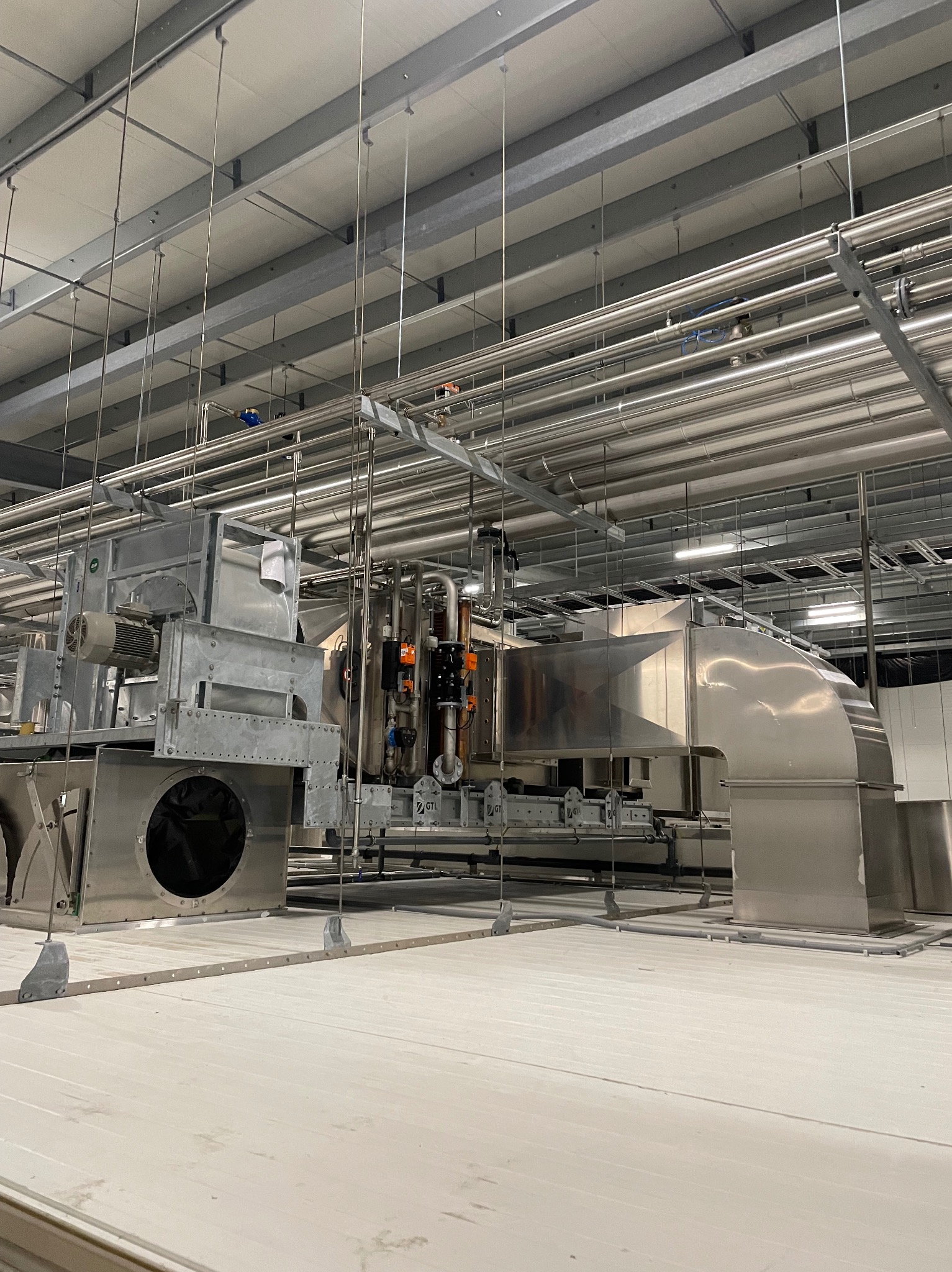 Srebrne kanały wentylacyjne i instalacja rekuperacji w dużej hali przemysłowej, widok z dołu na system rur podwieszonych pod sufitem.