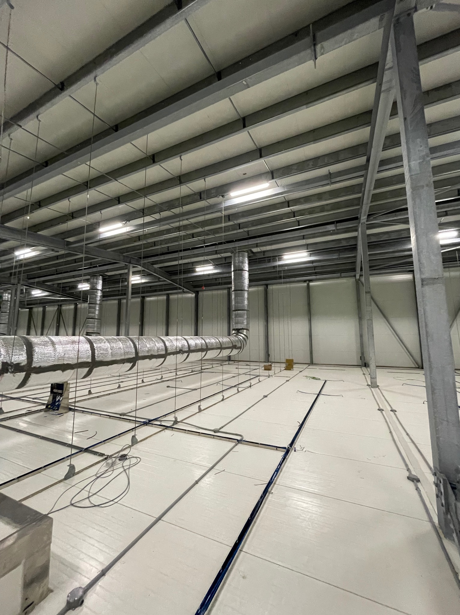 Wnętrze hali przemysłowej w trakcie montażu instalacji wentylacyjnej. Widoczne srebrne rury wentylacyjne podwieszone pod sufitem oraz elementy konstrukcji stalowej i oświetlenie.