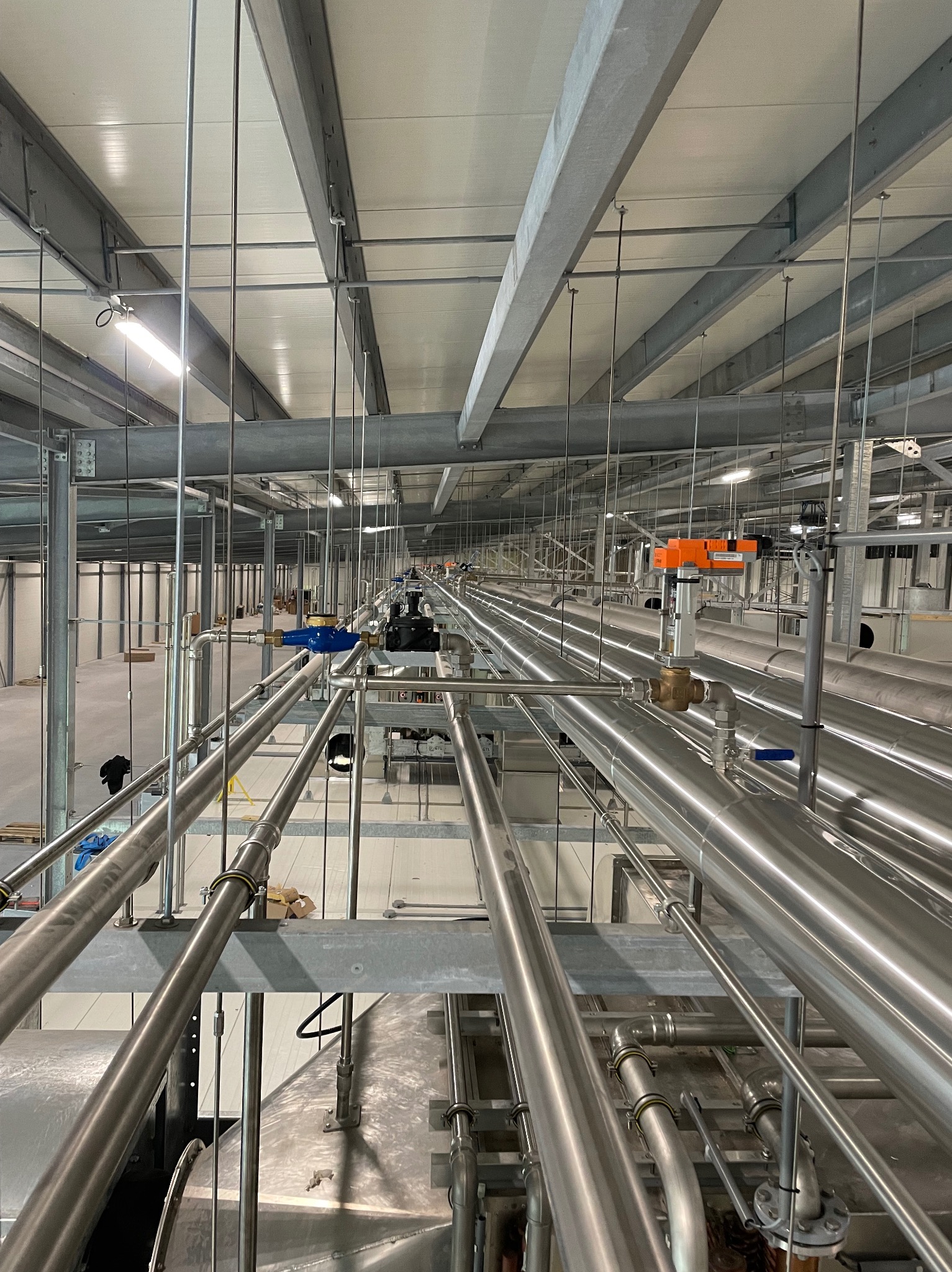 Rozbudowany system rur ze stali nierdzewnej pod sufitem hali przemysłowej, widoczne liczne zawory i łączenia, elementy wentylacji i konstrukcji wsporczej.