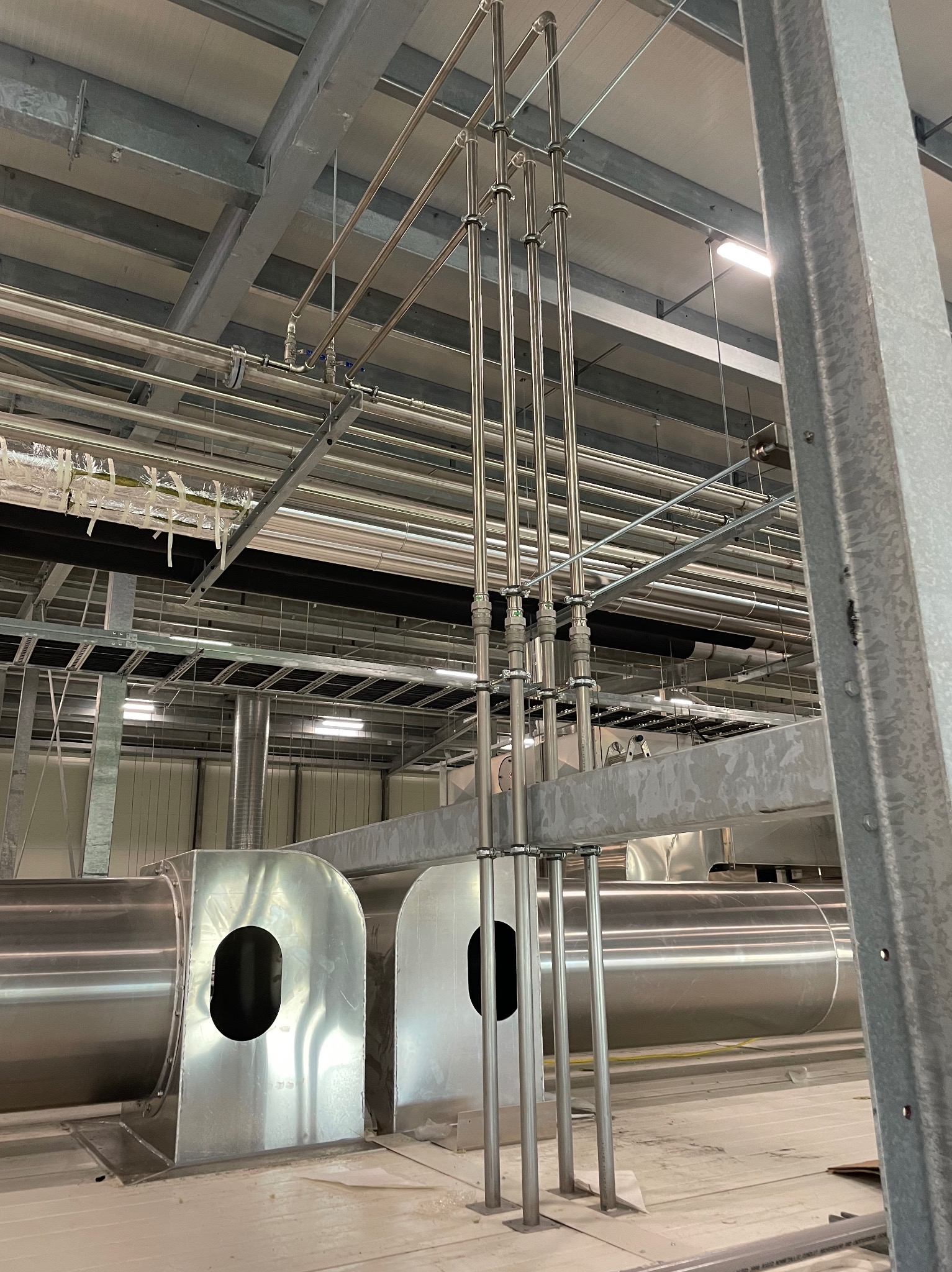 Metalowe rury wentylacyjne i hydrauliczne w hali przemysłowej, widoczne elementy mocujące i podwieszana konstrukcja sufitowa.