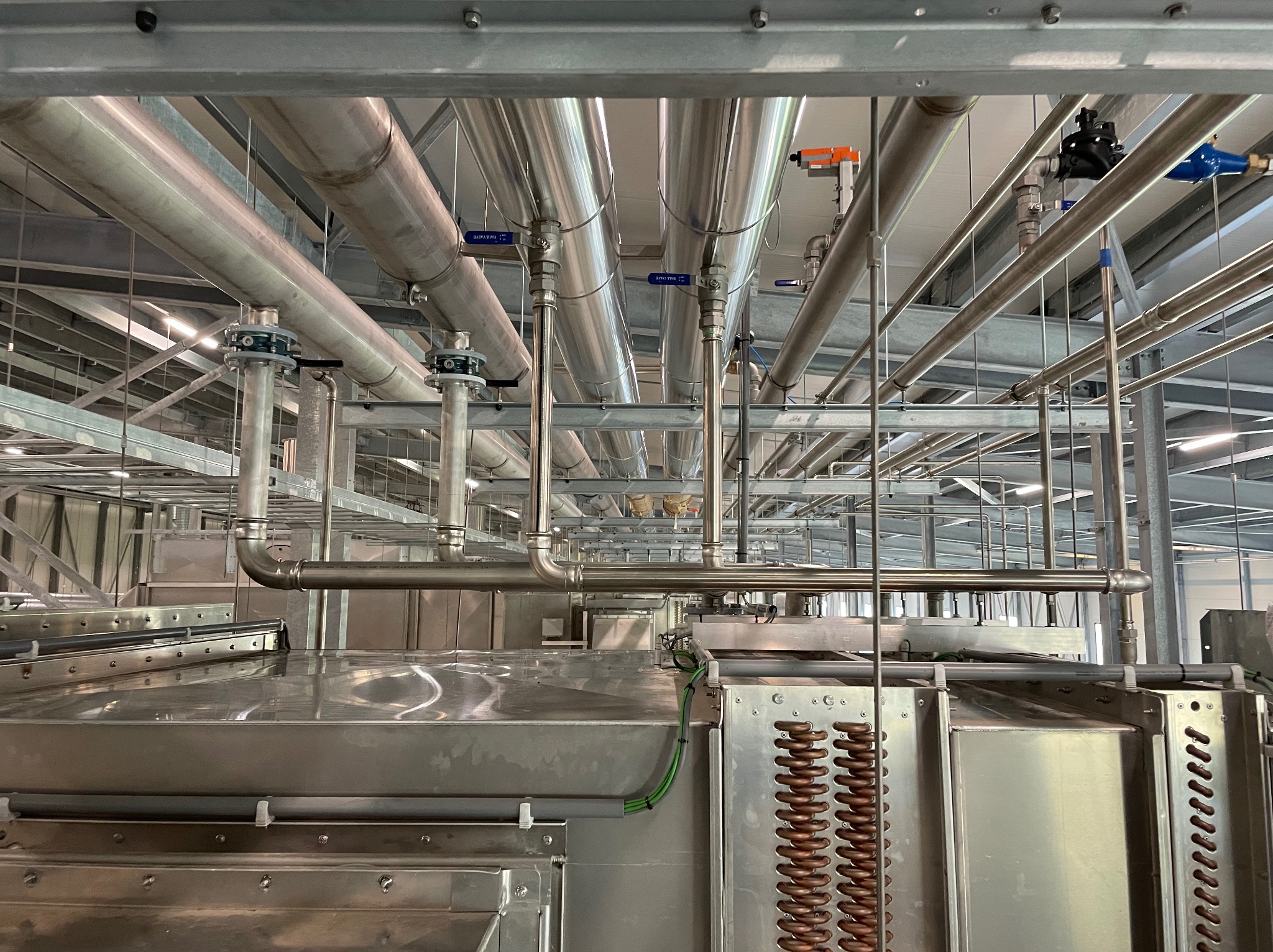 System rur ze stali nierdzewnej z zaworami i elementami mocującymi, prawdopodobnie w hali przemysłowej, widoczne również elementy chłodnicze.
