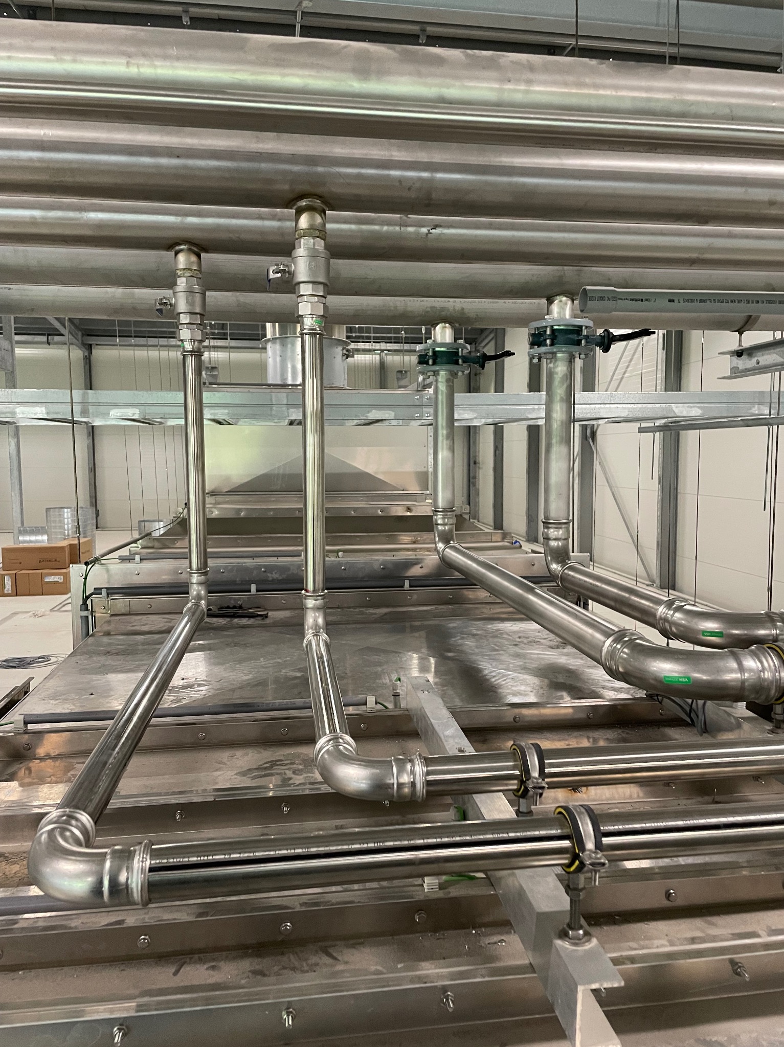 Szczegółowe ujęcie instalacji przemysłowej z rurami ze stali nierdzewnej, z widocznymi elementami mocującymi i zaworami, umieszczonej w jasnym pomieszczeniu przemysłowym.