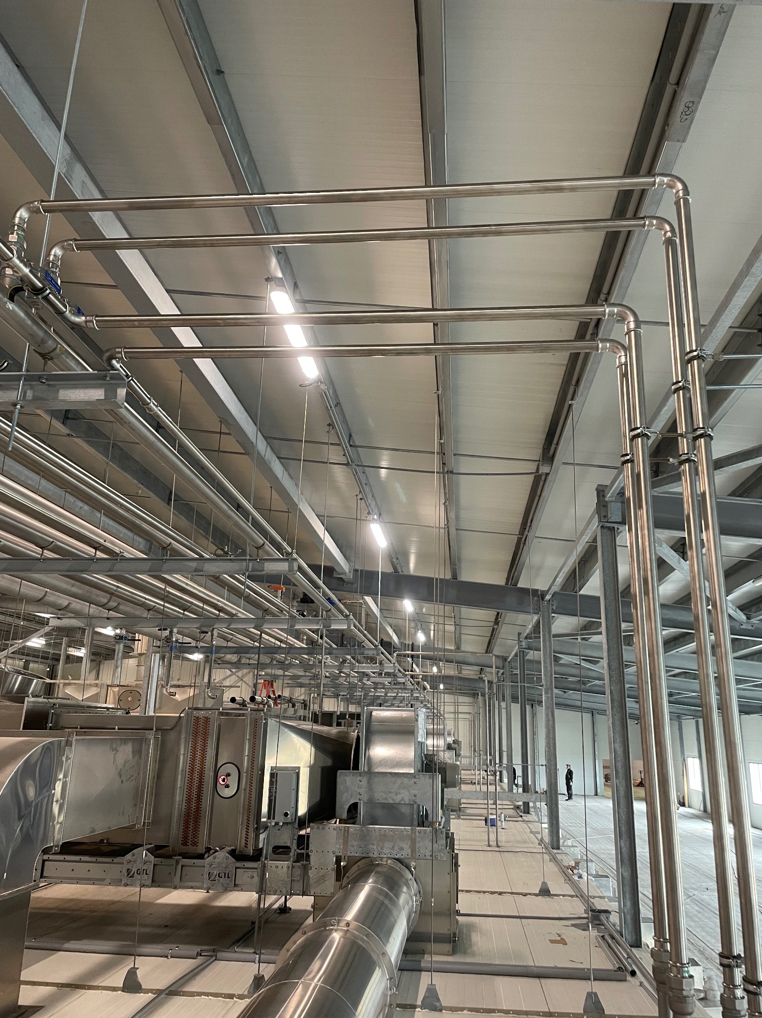 Widok na system rur i urządzeń wentylacyjnych w hali przemysłowej, z widocznymi elementami mocującymi i niewielką postacią ludzką w tle.