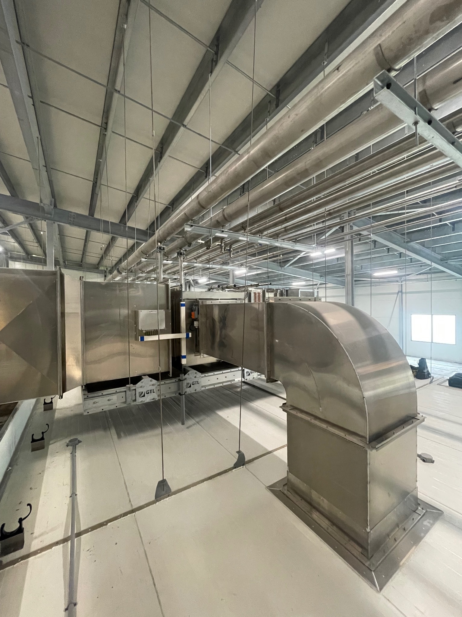 Srebrny kanał wentylacyjny z zaokrąglonym kolankiem, zamontowany na dachu hali przemysłowej z widoczną konstrukcją stalową pod sufitem i oknem w tle.