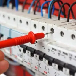 Pomiar napięcia w rozdzielnicy elektrycznej za pomocą miernika z czerwoną rączką, widoczne bezpieczniki i przewody.