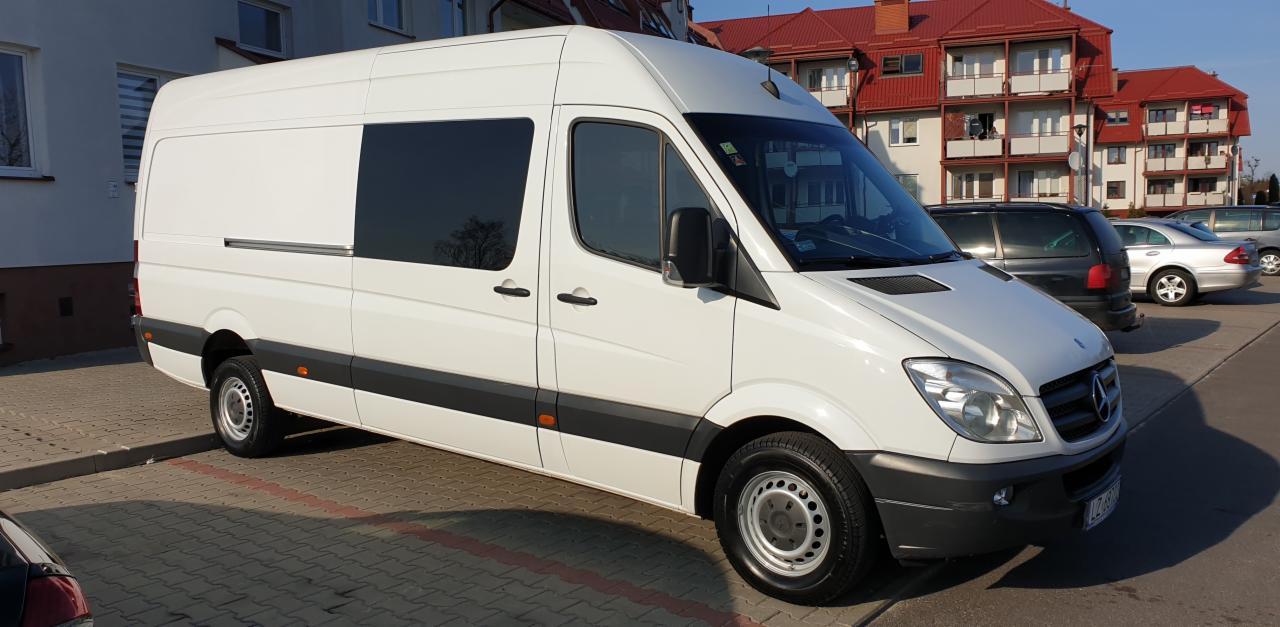 Biały samochód dostawczy Mercedes-Benz Sprinter z przyciemnionymi szybami, zaparkowany na kostce brukowej przed budynkiem mieszkalnym z czerwonym dachem.