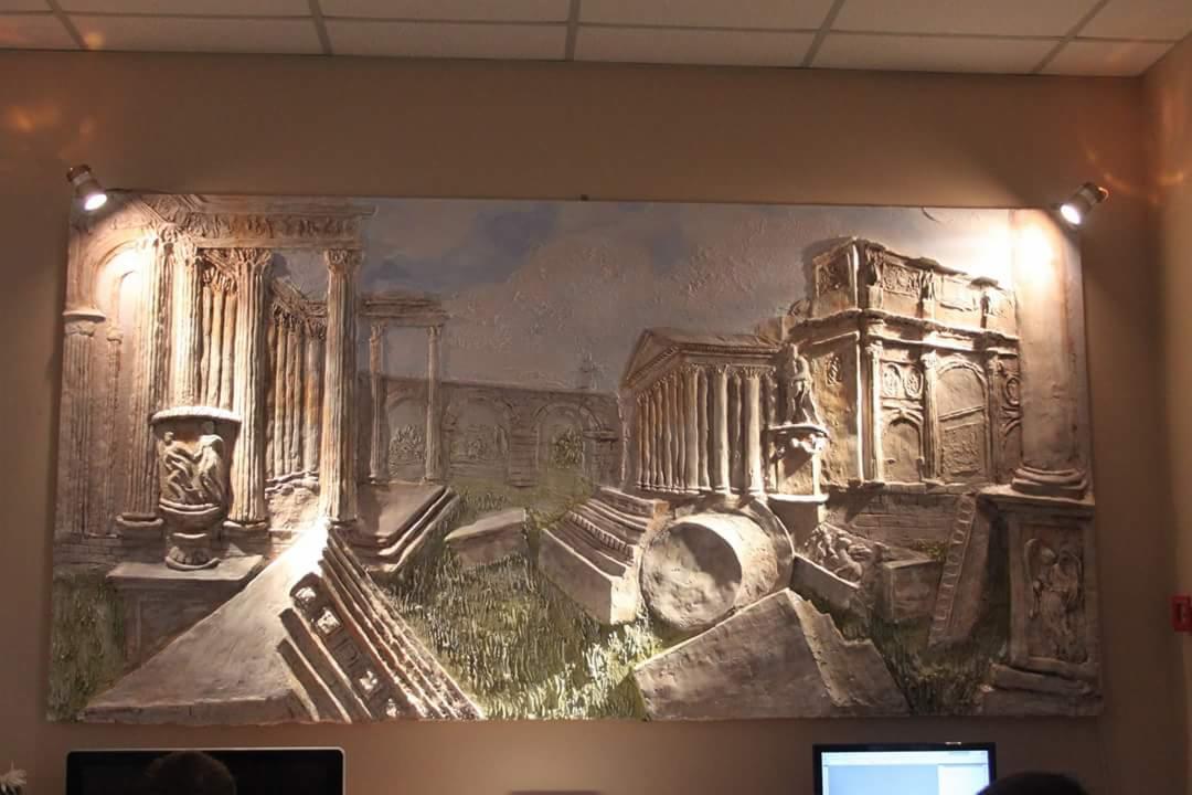 Relief ścienny przedstawiający ruinę starożytnego miasta z kolumnami i łukami, oświetlony dwoma reflektorami punktowymi.