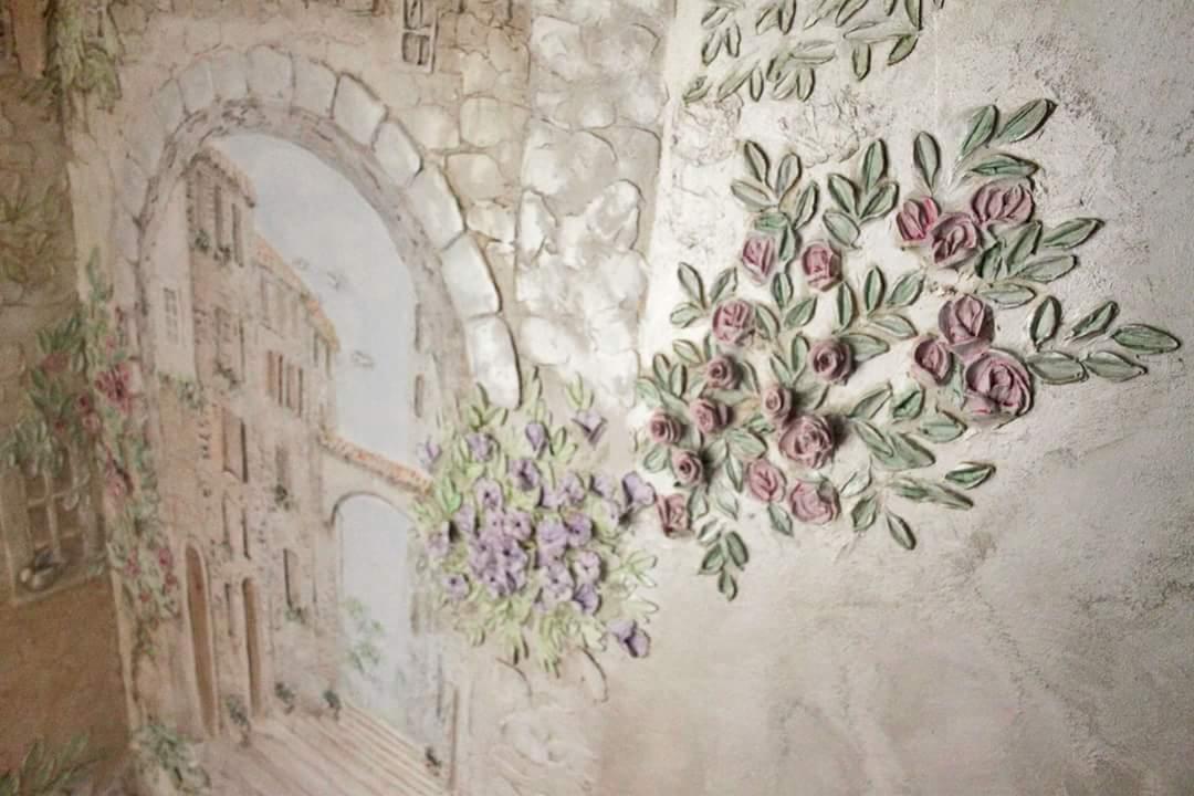 Dekoracyjna ściana z efektem 3D: szpachlowana imitacja kamiennego łuku z widokiem na włoską uliczkę, ozdobiona wypukłymi różami i innymi kwiatami w pastelowych kolorach.