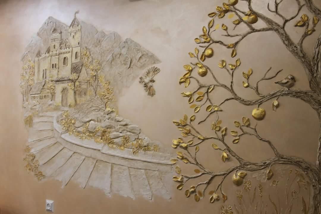 Dekoracyjna płaskorzeźba na ścianie przedstawiająca zamek w górach i drzewo z owocami, wykonana w odcieniach beżu i złota.