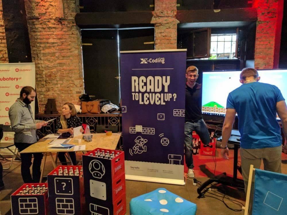 Konferencja X-Coding: Mężczyzna pozuje obok baneru z hasłem 'Ready to Level Up?', ekran z grą w tle, uczestnicy przy stoliku, skrzynki piwa z motywami gier.