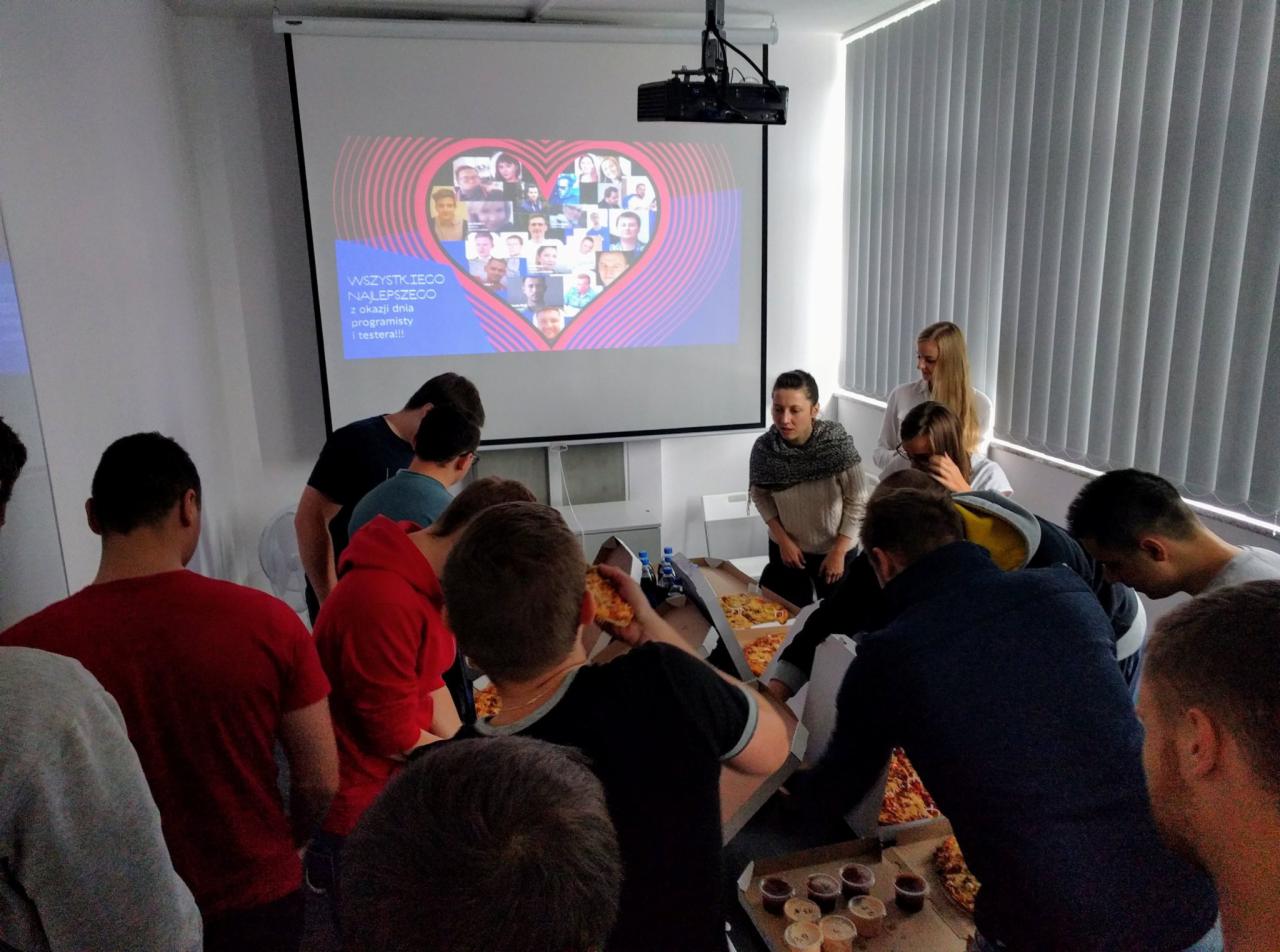 Grupa osób w biurze świętuje dzień programisty i testera, jedząc pizzę. Na ścianie wyświetlany jest projektor z kolażem zdjęć w kształcie serca.