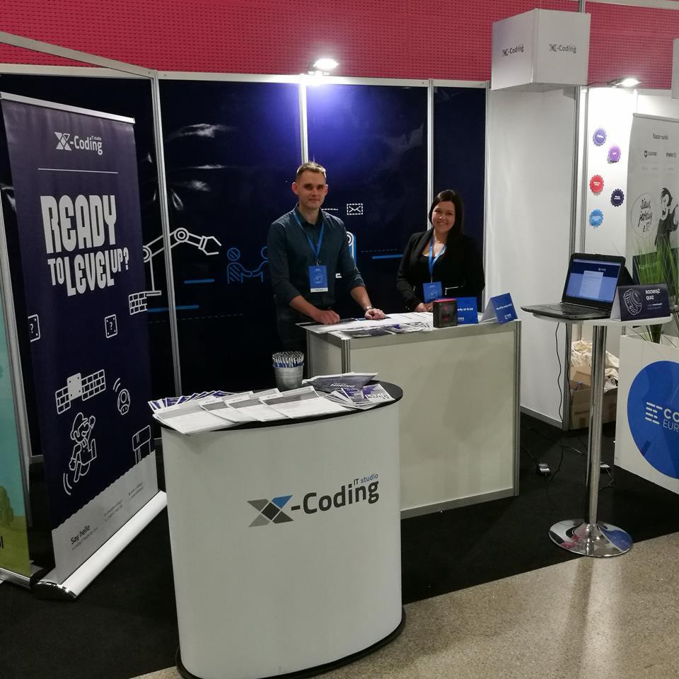 Stoisko targowe firmy IT z logo X-Coding, materiały informacyjne i dwoje pracowników za ladą w jasnych koszulach z identyfikatorami.