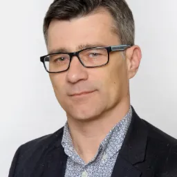 Portret mężczyzny w okularach i wzorzystej koszuli, ubrany w ciemną marynarkę, na jasnym tle.