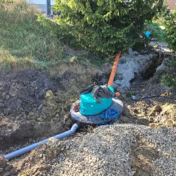 Zielony zbiornik przydomowej oczyszczalni częściowo zakopany w ziemi, widoczne rury, niebieski sznur i robocze rękawice na pokrywie, otoczenie wykopane w ziemi z kamieniami i trawą.