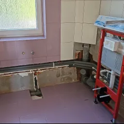 Remont łazienki w trakcie: widoczne rury instalacyjne, stelaż podtynkowy WC z opakowaniem odpływu liniowego, częściowo skute płytki, okno z widokiem na zieleń.