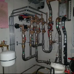 Skomplikowana instalacja hydrauliczna z rur miedzianych, stalowych i plastikowych, z widocznymi manometrami, zaworami i zbiornikiem wyrównawczym, zamontowana na surowej ścianie w piwnicy.