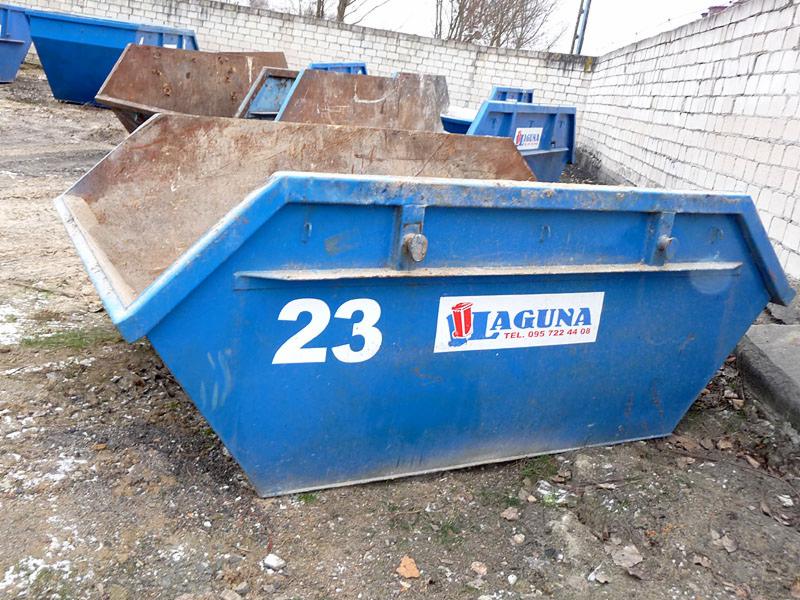 Stos kontenerów na gruz, jeden niebieski z numerem 23 i logo firmy Laguna, pozostałe rdzewiejące, ustawione na ziemi przy ceglanym murze.