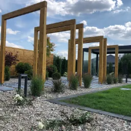 Nowoczesna aranżacja ogrodu z drewnianą konstrukcją pergoli, ścieżką z płyt kamiennych i żwirowym podłożem, uzupełniona o roślinność ozdobną i oświetlenie ogrodowe, z widoczną w tle drewnianą ścianą.