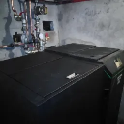 Czarny piec z widoczną instalacją hydrauliczną w piwnicy, rury miedziane i stalowe, szare ściany betonowe.