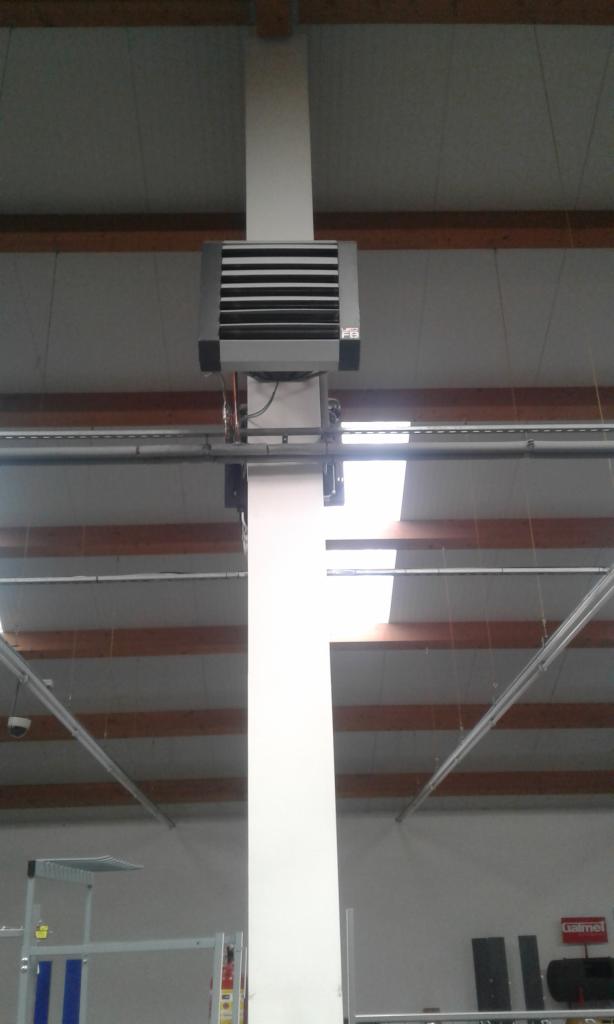 Szara nagrzewnica powietrza zamontowana na białej kolumnie w hali przemysłowej z widoczną konstrukcją dachu i oświetleniem.