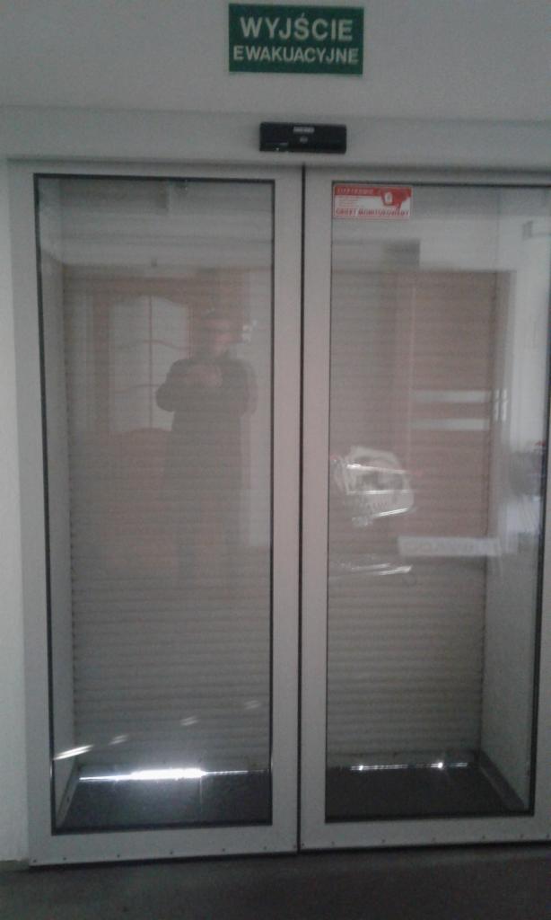 Automatyczne, dwuskrzydłowe drzwi szklane z napisem 'Wyjście Ewakuacyjne' u góry, wyposażone w rolety wewnętrzne, widoczne odbicie osoby w szybie.