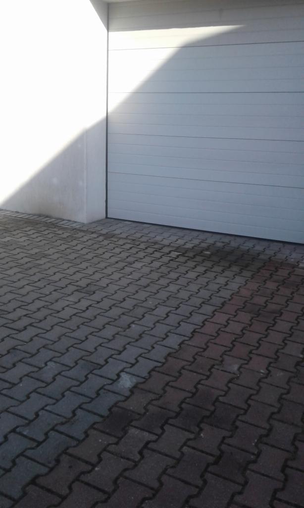 Zamknięta, segmentowa brama garażowa w kolorze jasnoszarym, z widocznym fragmentem elewacji budynku oświetlonym padającym światłem słonecznym oraz kostką brukową przed bramą.