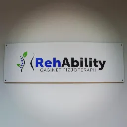 Szyld gabinetu fizjoterapii z napisem 'RehAbility Gabinet Fizjoterapii' na białym tle, przymocowany do ściany. Logo zawiera stylizowany motyw roślinny z lewej strony.