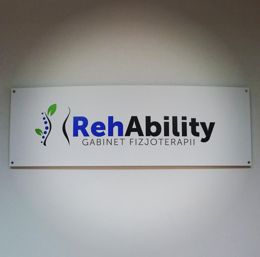 Szyld gabinetu fizjoterapii z napisem 'RehAbility Gabinet Fizjoterapii' na białym tle, przymocowany do ściany. Logo zawiera stylizowany motyw roślinny z lewej strony.