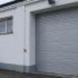 Szara, metalowa osłona przeciwsłoneczna okna lub drzwi w budynku o białej fasadzie z widocznymi śladami zabrudzeń i donicą z rośliną po lewej stronie.