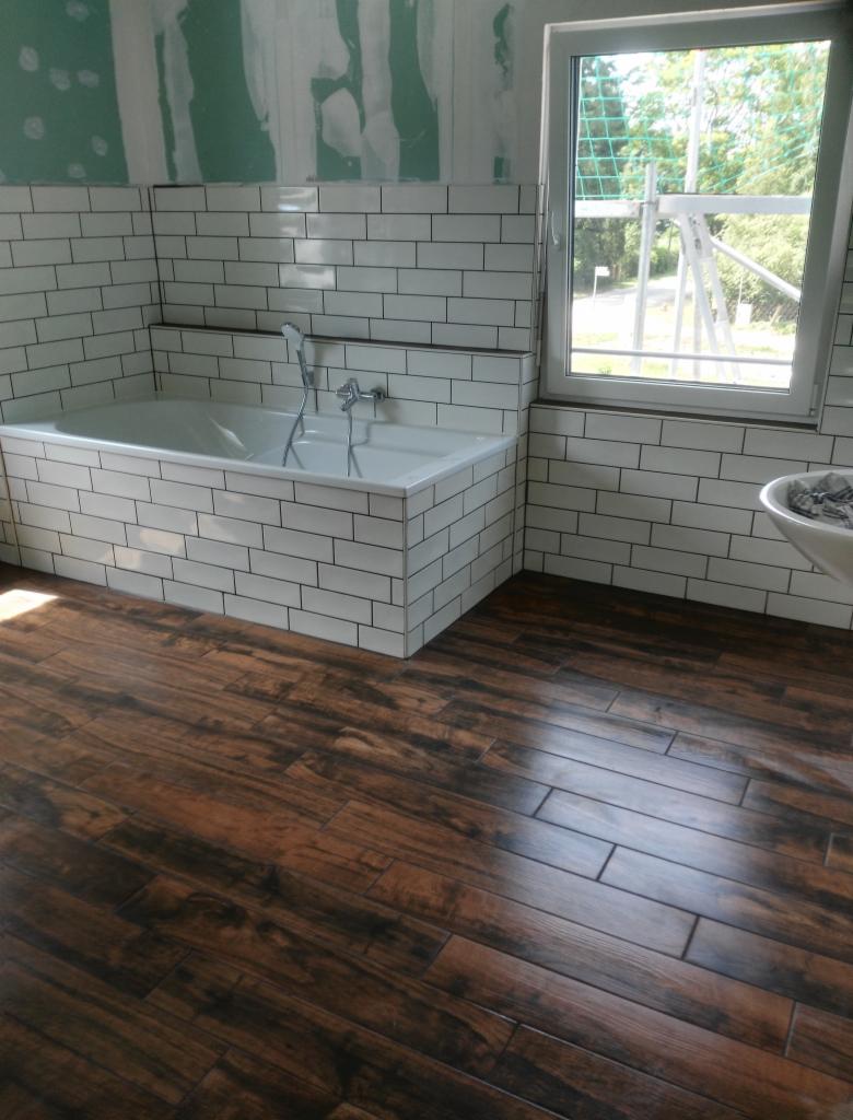 Wnętrze łazienki w trakcie prac wykończeniowych: białe, prostokątne płytki na ścianach, drewnopodobna podłoga, wanna obudowana płytkami, okno z widokiem na zewnątrz, fragment ściany z zieloną płytą...