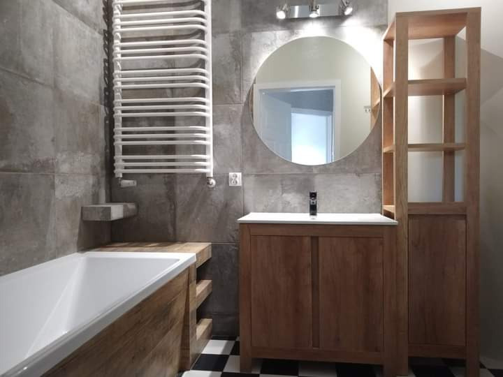 Wykończona łazienka z szarymi płytkami, drewnianą zabudową wanny i umywalki, okrągłym lustrem z podświetleniem oraz grzejnikiem drabinkowym.